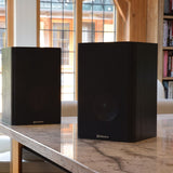 Adastra AB-5 passive bookshelf speakers in matt black woodgrain finish, displayed as a pair on a marble surface near a window.