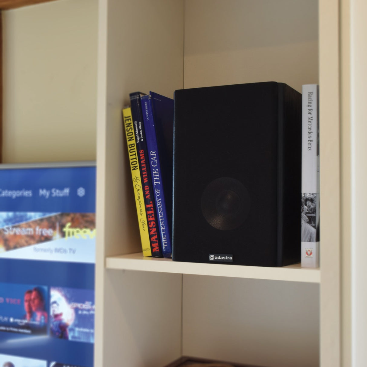 Adastra AB-5 matt black bookshelf speaker on a cream shelf beside motorsport books, next to a TV showing a streaming menu.