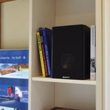 Adastra AB-5 matt black bookshelf speaker on a cream shelf beside motorsport books, next to a TV showing a streaming menu.