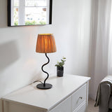 Endon Danemont table lamp with natural raffia tapered shade and wavy matt black metal stem, on a white dresser beside a potted plant in a bedroom.
