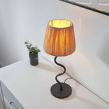 Endon Danemont table lamp with a natural raffia tapered shade on a wavy matt black metal stem and round base, lit on a white sideboard.