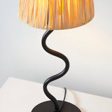 Endon Danemont table lamp with a natural raffia tapered shade and matt black wavy metal stem, illuminated on a white surface.