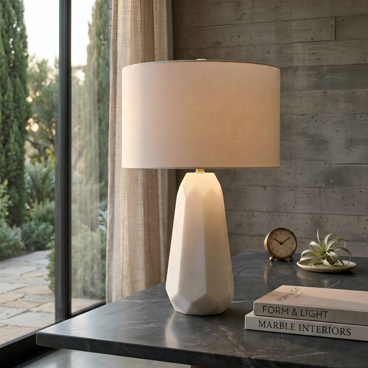 The Averon 1 Light Faceted Alabaster Table Lamp in Brass & Soft White sits on a dark marble desk by a large window, beside stacked books, a clock, a small plant, and beige curtains with greenery outside.