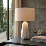 The Averon 1 Light Faceted Alabaster Table Lamp in Brass & Soft White sits on a dark marble desk by a large window, beside stacked books, a clock, a small plant, and beige curtains with greenery outside.