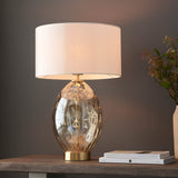 Endon Draycott table lamp with dimpled amber glass base, satin brass fittings and white fabric drum shade, lit on a wooden console beside books and flowers.