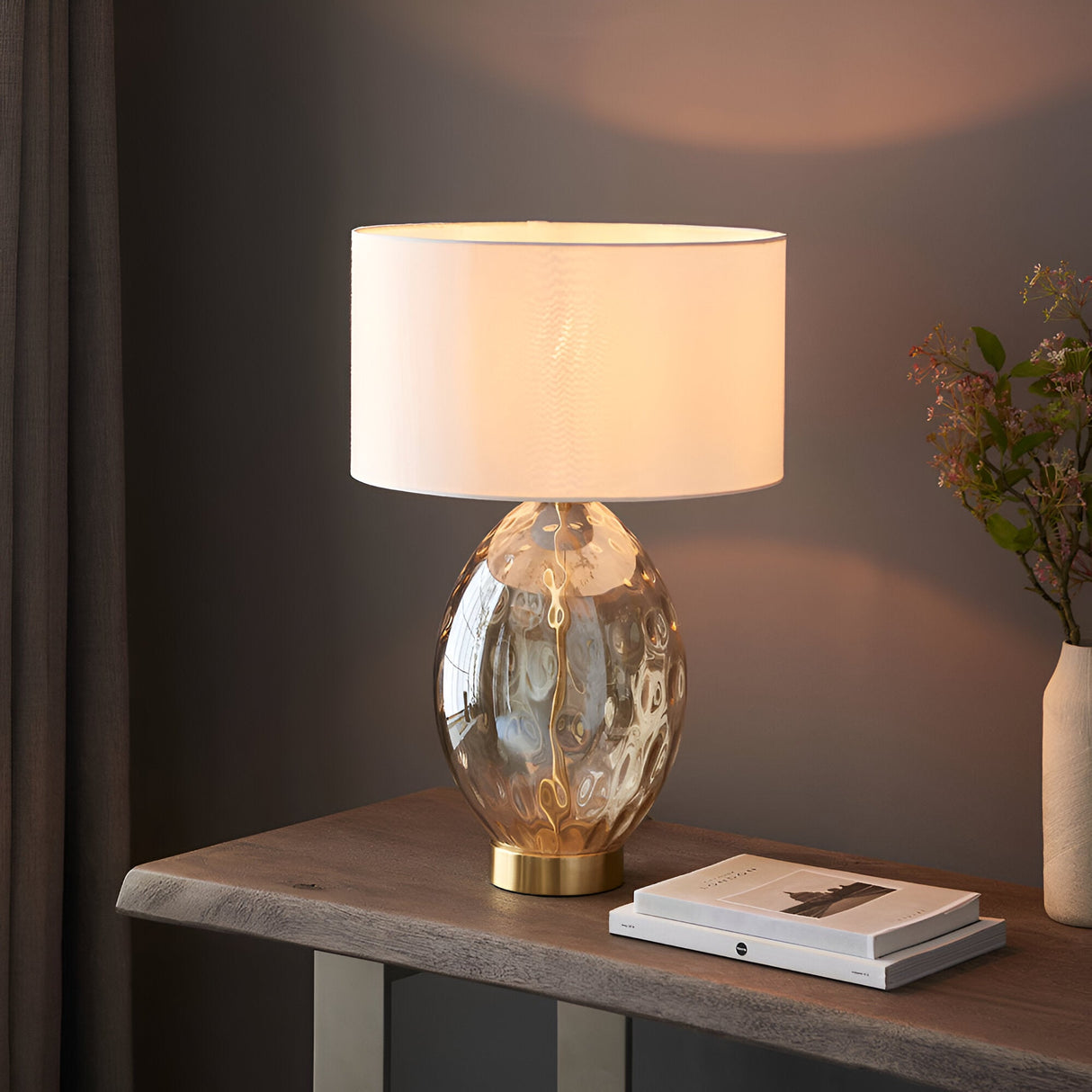 Endon Draycott table lamp with dimpled amber glass base, satin brass fitting and white fabric drum shade, lit on a wood console beside books.