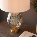Endon Draycott dimpled glass table lamp with satin brass base and vintage white fabric drum shade on a wooden bedside beside pink blossoms.
