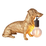 Bevworth Table Lamp shaped as a gold resin dachshund holding a black E27 lamp fitting with exposed filament bulb in its mouth, on a plain white background.
