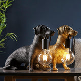 Bevworth Table Lamp pair in gold and black resin dachshund shapes, each holding an exposed E27 filament bulb, on a dark table setting.