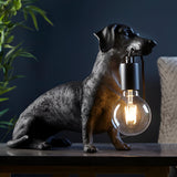 Bevworth Table Lamp in Black: a resin dachshund figure holding an E27 filament bulb in its mouth, sitting on a dark tabletop.