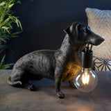 Bevworth Table Lamp in black resin, shaped as a seated dachshund holding an E27 filament bulb in its mouth, set against a dark room backdrop.