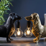 Bevworth Table Lamp in black and vintage gold resin dachshund shapes, each holding an exposed E27 filament bulb, set against a dark background.