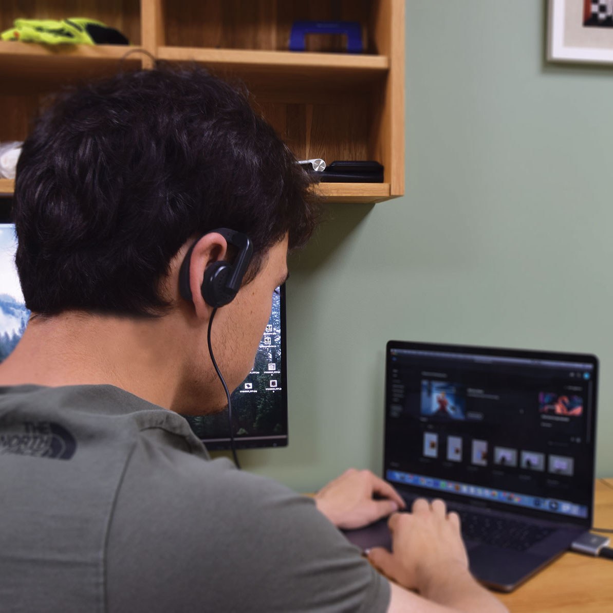Man at desk wearing the AVSL AV:Link ME22 mono earphone with adjustable ear-hook, cable trailing to a laptop and monitor in a home workspace.