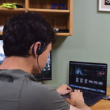 Man at desk wearing the AVSL AV:Link ME22 mono earphone with adjustable ear-hook, cable trailing to a laptop and monitor in a home workspace.