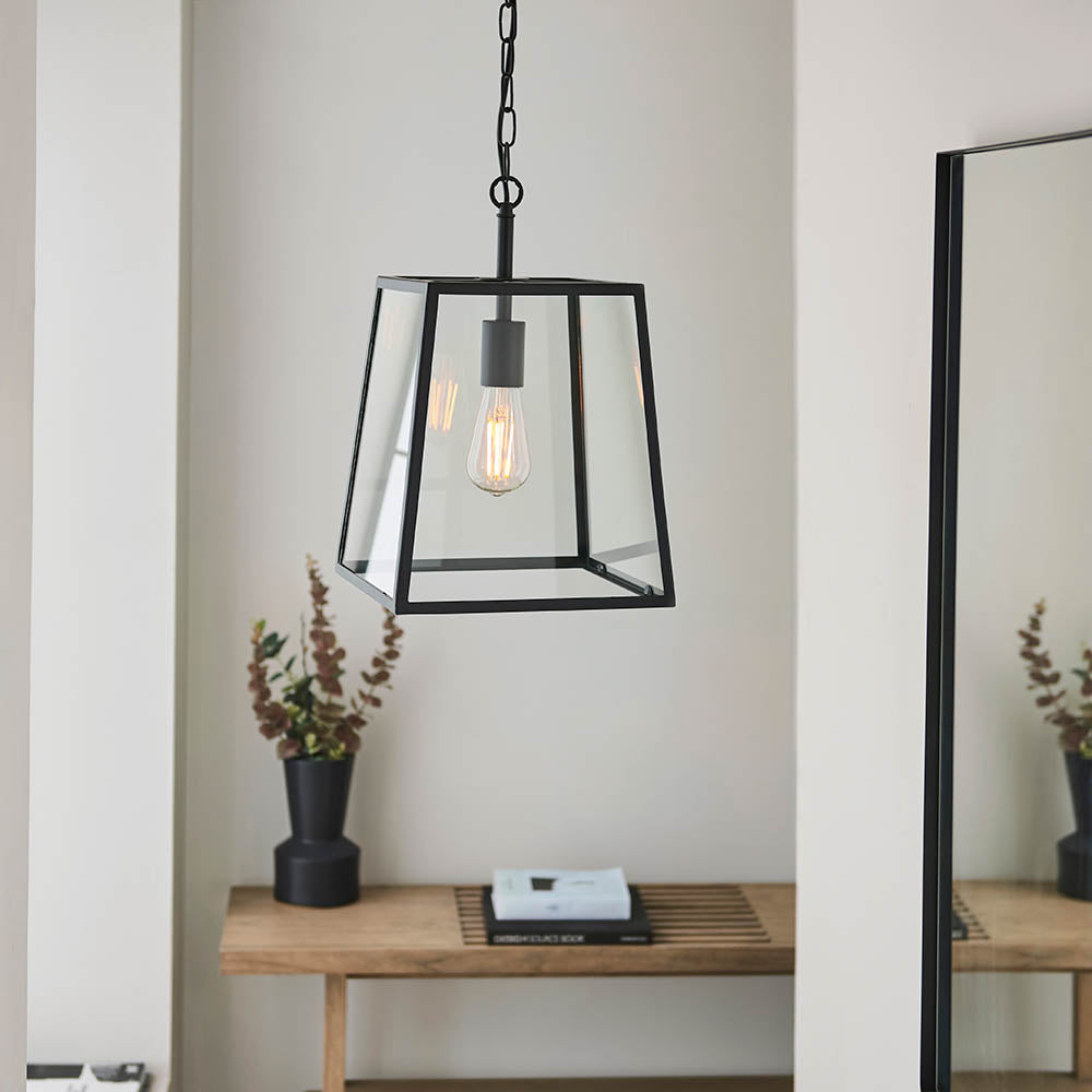 Brenton Lantern Pendant in black hangs from a chain, its tapered glass-panelled frame revealing an exposed filament bulb above a wooden console table.