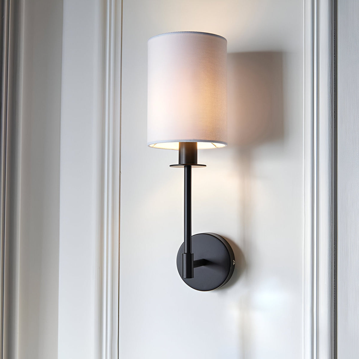 Endon Cliffhurst wall light with a slim matt black upright and round backplate topped by a vintage white cylinder shade, mounted on a panelled wall.