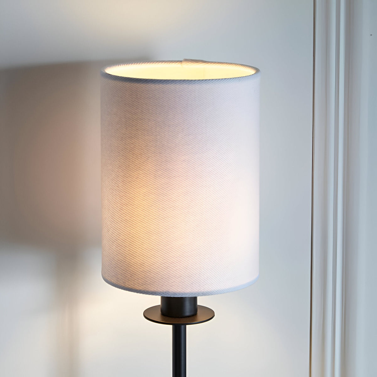 Endon Cliffhurst wall light with slim matt black upright and vintage white cylinder fabric shade, illuminated against a pale wall.