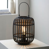 Endon Bracken table lamp in matt black with an open bamboo cage, arched handle and lit E27 filament bulb, shown on a white side table.