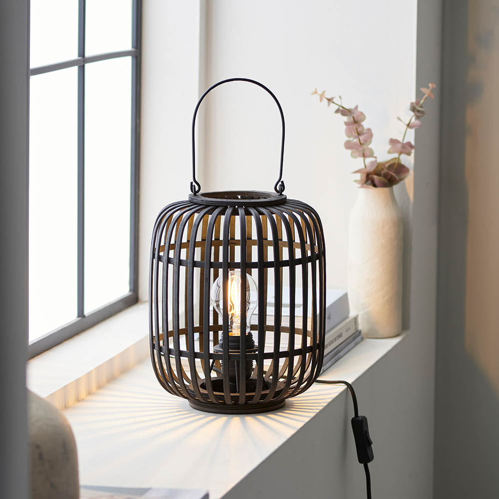 Endon Bracken table lamp in black with an open bamboo cage, arched handle and E27 filament bulb, lit on a windowsill beside a vase of eucalyptus.