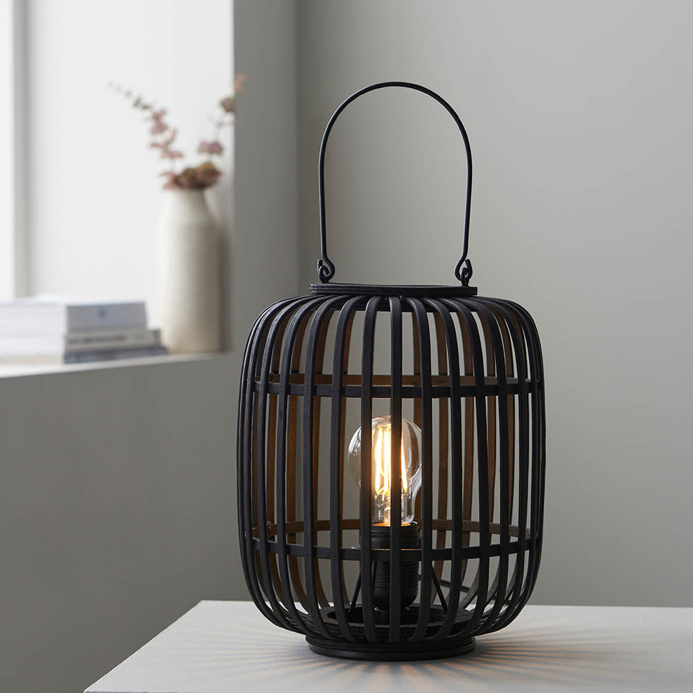 Endon Bracken table lamp in black with open bamboo cage, arched handle and visible filament bulb, shown lit on a side table by a bright window.