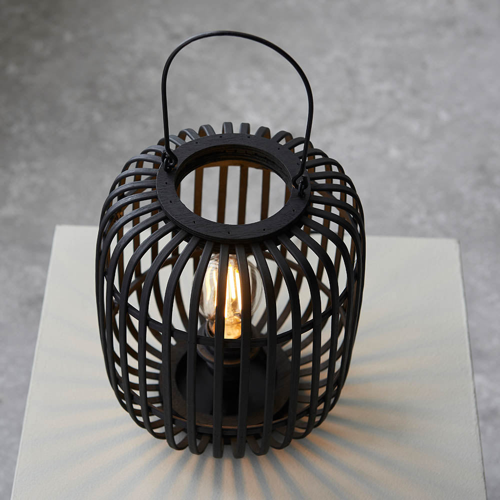 Endon Bracken table lamp in black with an open bamboo cage, decorative handle and visible filament E27 bulb, shown lit on a pale plinth.
