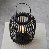 Endon Bracken table lamp in black with an open bamboo cage, decorative handle and visible filament E27 bulb, shown lit on a pale plinth.