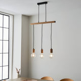 Cranbury 3 Light Linear Pendant in black and natural wood, with exposed filament bulbs hanging above a dining table by a large window.