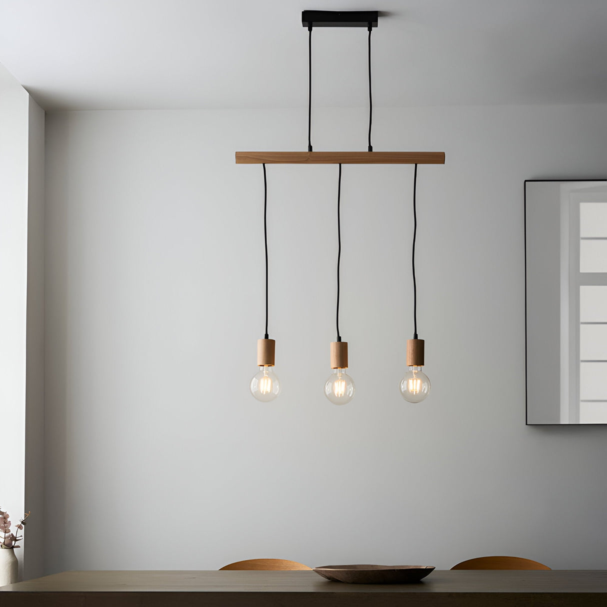 Cranbury 3 Light Linear Pendant in black and natural wood, hung above a dining table with exposed filament bulbs, set against a plain white wall.