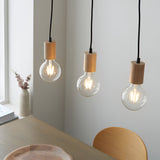 Cranbury 3 Light Linear Pendant in black and natural wood, three exposed filament bulbs hanging at staggered heights above a dining table.