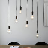 Endon Cliffwell 6 light linear pendant with exposed filament bulbs on black flex and anthracite lampholders, hung over a dining table by a pale wall.