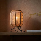 Endon Daneshaw table lamp with a lantern-shaped natural linen and bamboo shade on a matt black metal base, lit on a wooden shelf against a warm wall.