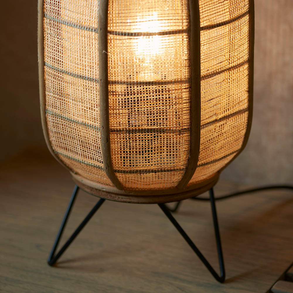 Endon Daneshaw table lamp lit on a wooden surface, showing its woven natural linen and bamboo shade above a matt black metal tripod base.