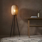 Daneshaw Floor Lamp with woven natural linen shade on bamboo frame and black tripod base, lit in a room beside a wooden table.