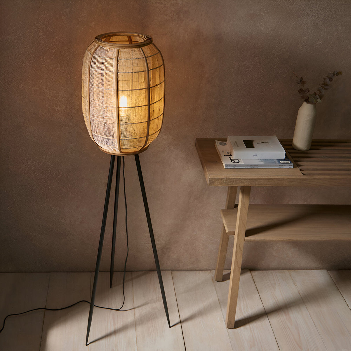 Daneshaw Floor Lamp with natural linen and bamboo cylindrical shade on a matt black tripod base, glowing warmly beside a wooden side table.
