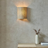 Endon Salcombe wall light with woven natural seagrass half-cylinder shade glowing on a textured plaster wall above a wooden bench with a vase.