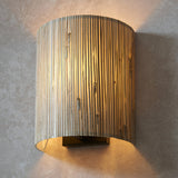 Endon Salcombe wall light with a woven natural seagrass half-cylinder shade on matt black metalwork, casting a warm glow against a stone wall.