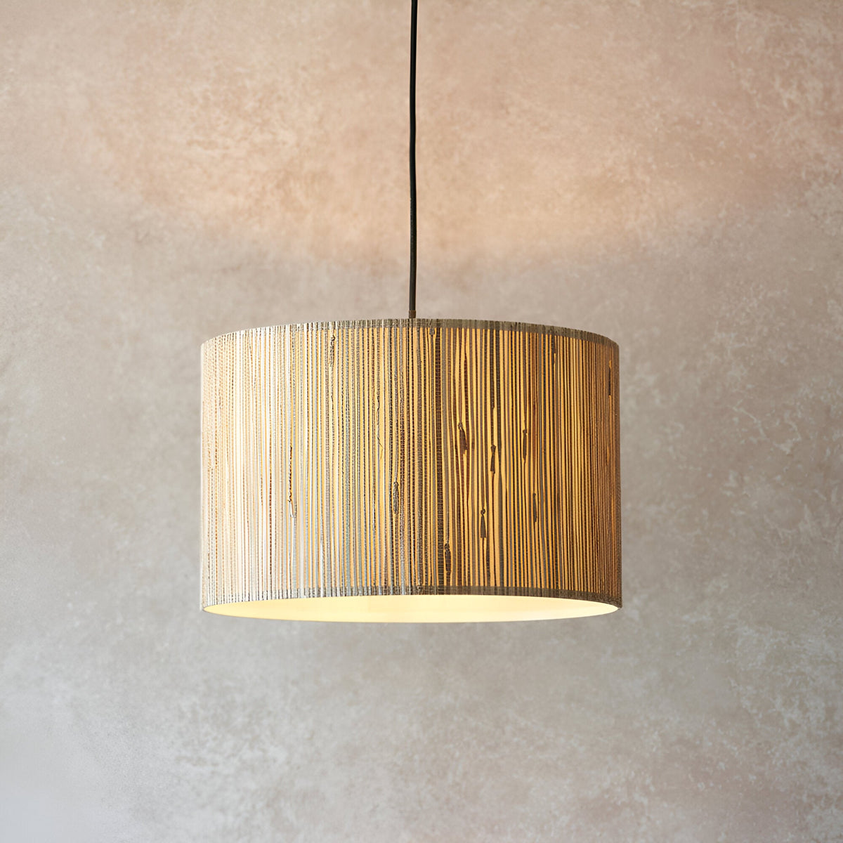 Salcombe Pendant Light in black and natural, a drum-shaped fixture with woven seagrass shade on a black cable, shown lit against a plain neutral wall.