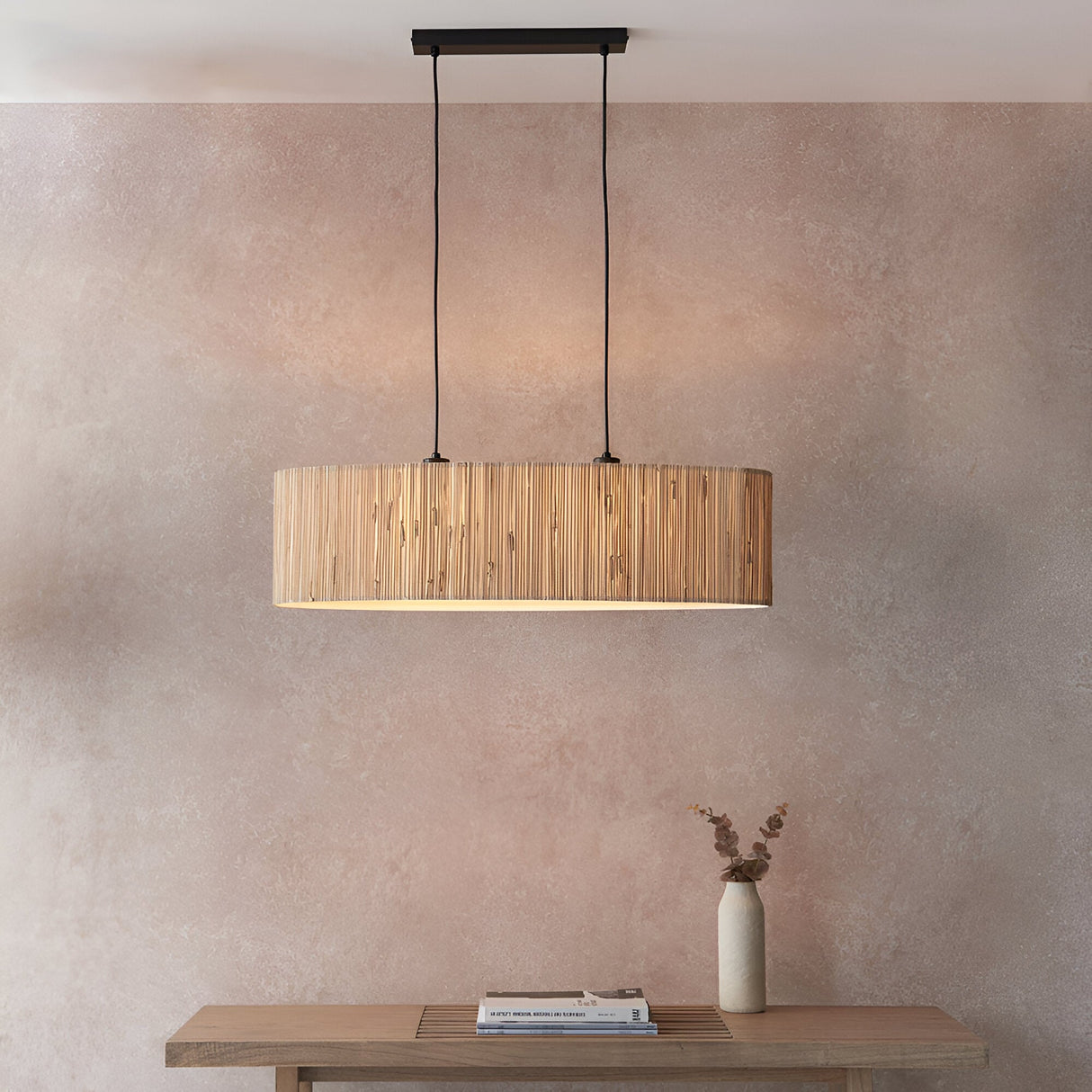 Endon Salcombe 2 light linear pendant with woven natural seagrass oval shade and matt black cables, hanging above a wooden console against a warm pink wall.
