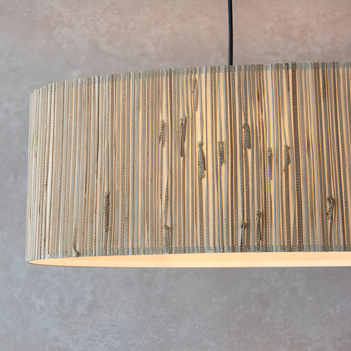 Endon Salcombe linear pendant close-up showing the woven natural seagrass oval shade with visible grain and matt black cable against a light grey wall.