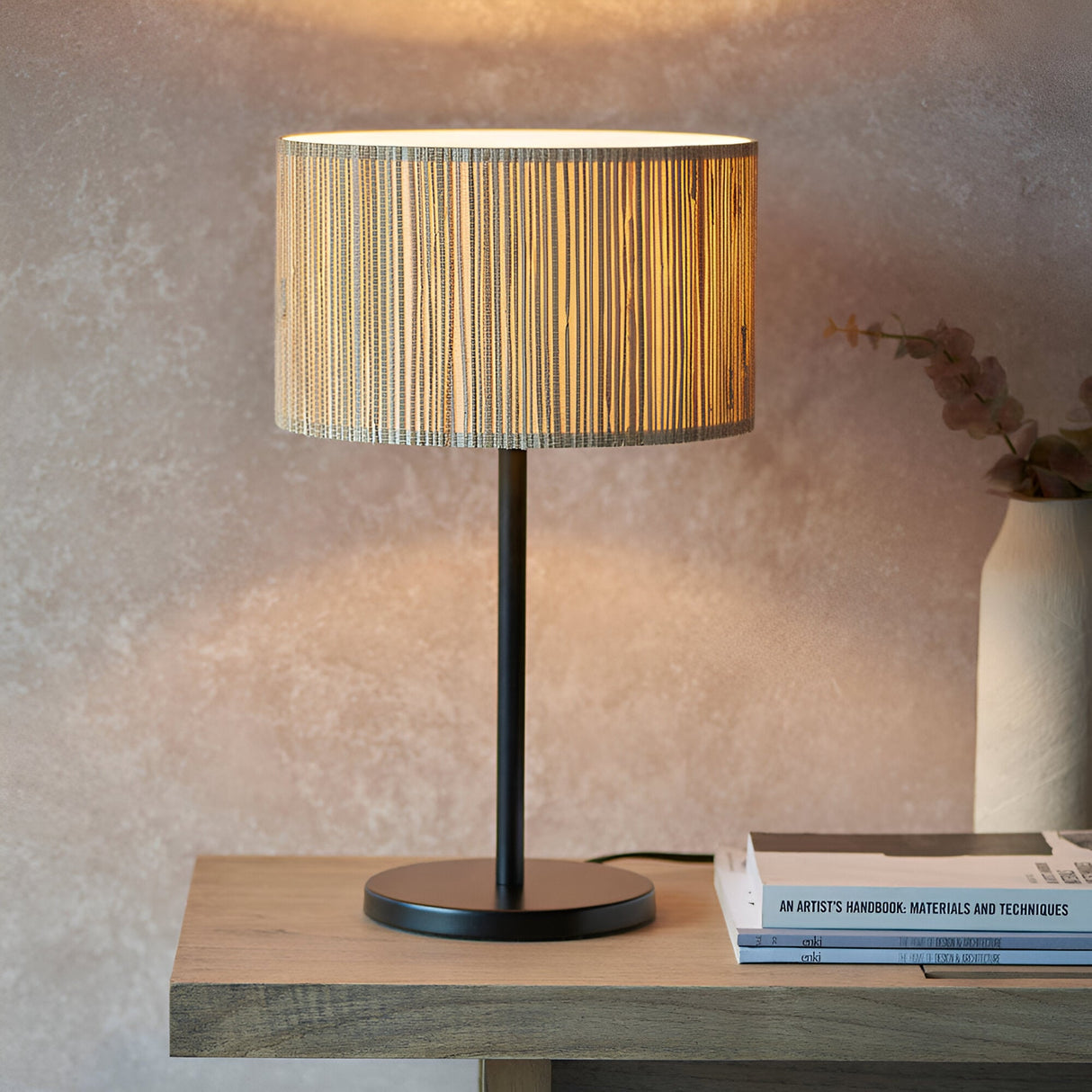 Salcombe Table Lamp with woven seagrass drum shade on a matt black metal base, styled on a wooden table beside stacked books.