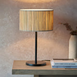 Salcombe Table Lamp with woven seagrass drum shade on a matt black metal base, styled on a wooden table beside stacked books.