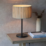 Salcombe Table Lamp with woven seagrass drum shade on a matt black metal base and stem, shown lit on a wooden table beside books and a vase.