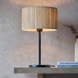 Salcombe Table Lamp with woven seagrass drum shade on a matt black metal base and stem, shown lit on a wooden console table.