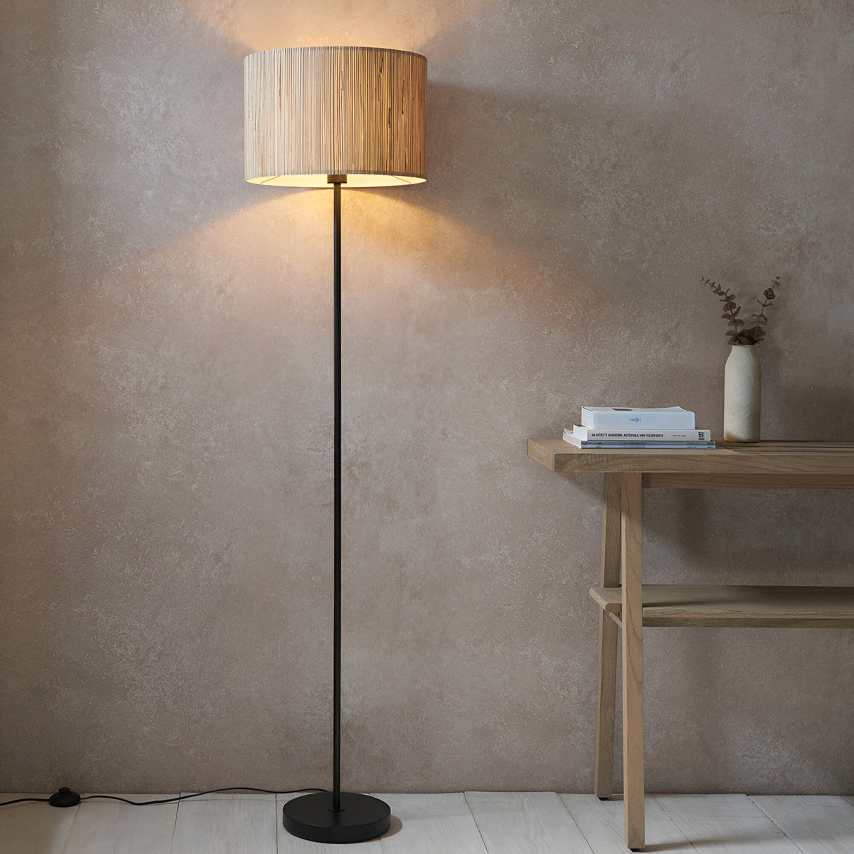 Endon Salcombe floor lamp with slim matt black stem and woven natural seagrass drum shade, lit beside a wooden console table against a plaster wall.