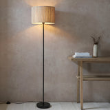 Endon Salcombe floor lamp with slim matt black stem and woven natural seagrass drum shade, lit beside a wooden console table against a plaster wall.