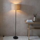 Endon Salcombe floor lamp with slim matt black stem and woven natural seagrass drum shade, lit beside a wooden slatted console table in a living space.