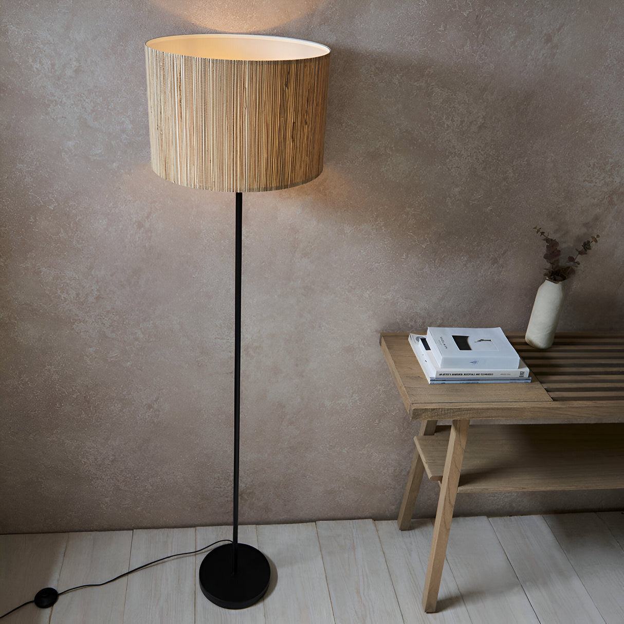 Endon Salcombe floor lamp with slim matt black stem and woven natural seagrass drum shade, lit beside a wooden bench in a warm neutral room.