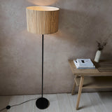 Endon Salcombe floor lamp with slim matt black stem and woven natural seagrass drum shade, lit beside a wooden bench in a warm neutral room.