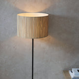 Endon Salcombe floor lamp with slim matt black stem and woven natural seagrass drum shade, lit beside a textured plaster wall.