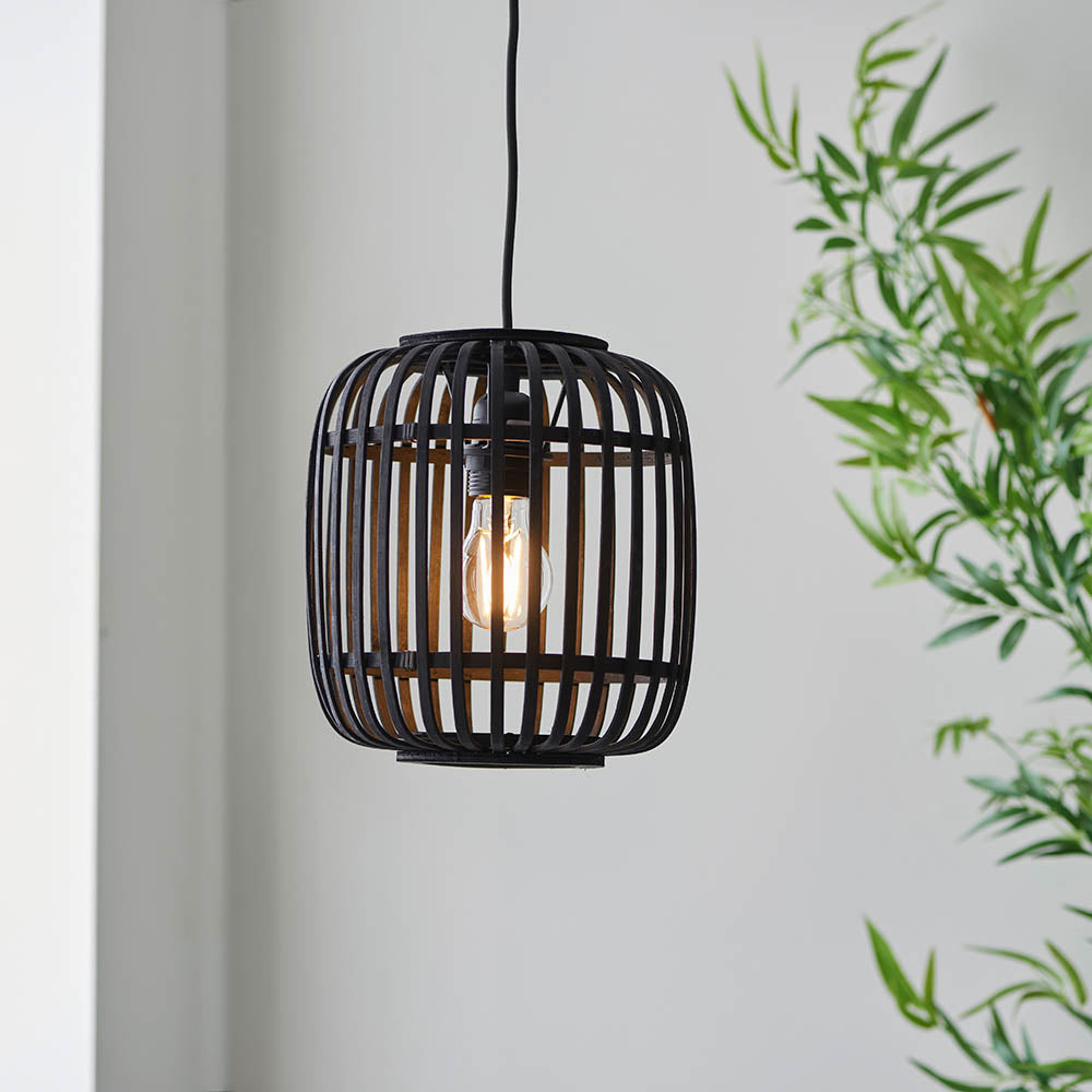 Bracken Pendant Light in black bamboo cage design with visible E27 filament bulb, hanging from black flex against a light wall near a green plant.